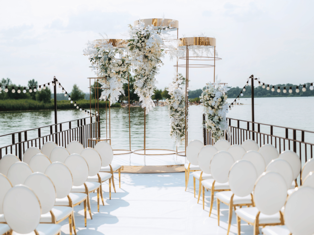 Outdoor wedding setup by the lake with rows of white chairs facing a floral archway and a string of lights, creating an elegant ceremony space.